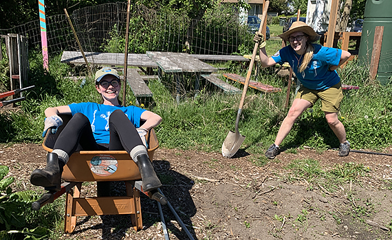 One woman lounges in a wheelbarrow while another poses with a shovel at an urban farm