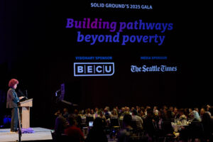 Woman with a red afro wearing white shirt and dark jacket speaks at a podium on a stage to a filled ballroom. The words "Building pathways beyond poverty" is projected in blue and purple on the wall.