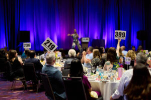 People seated around tables in a purple-hued ballroom thrust bid cards in the air as someone out-of-focus on stage gestures.
