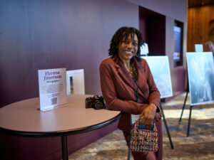 A young woman in a brown suit smiles and leans against a round table in a convention center lobby.