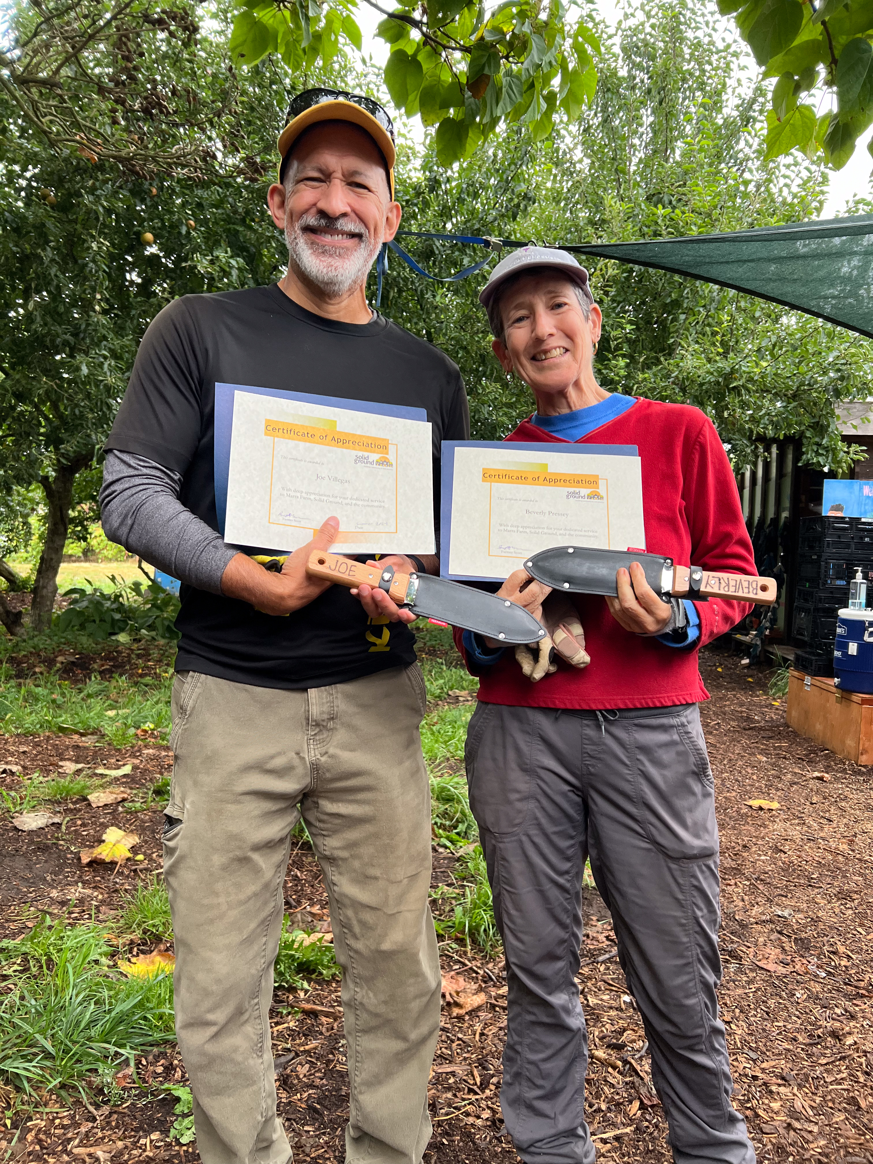 Two people wearing caps and work clothes pose smiling under a plum tree, holding gardening tools and certificates of appreciation.