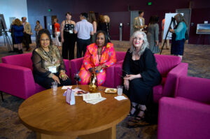 Three women sit in pink plush chairs around a table.