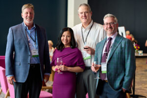 A group of people pose in dressy attire, some of them holding glasses of wine.