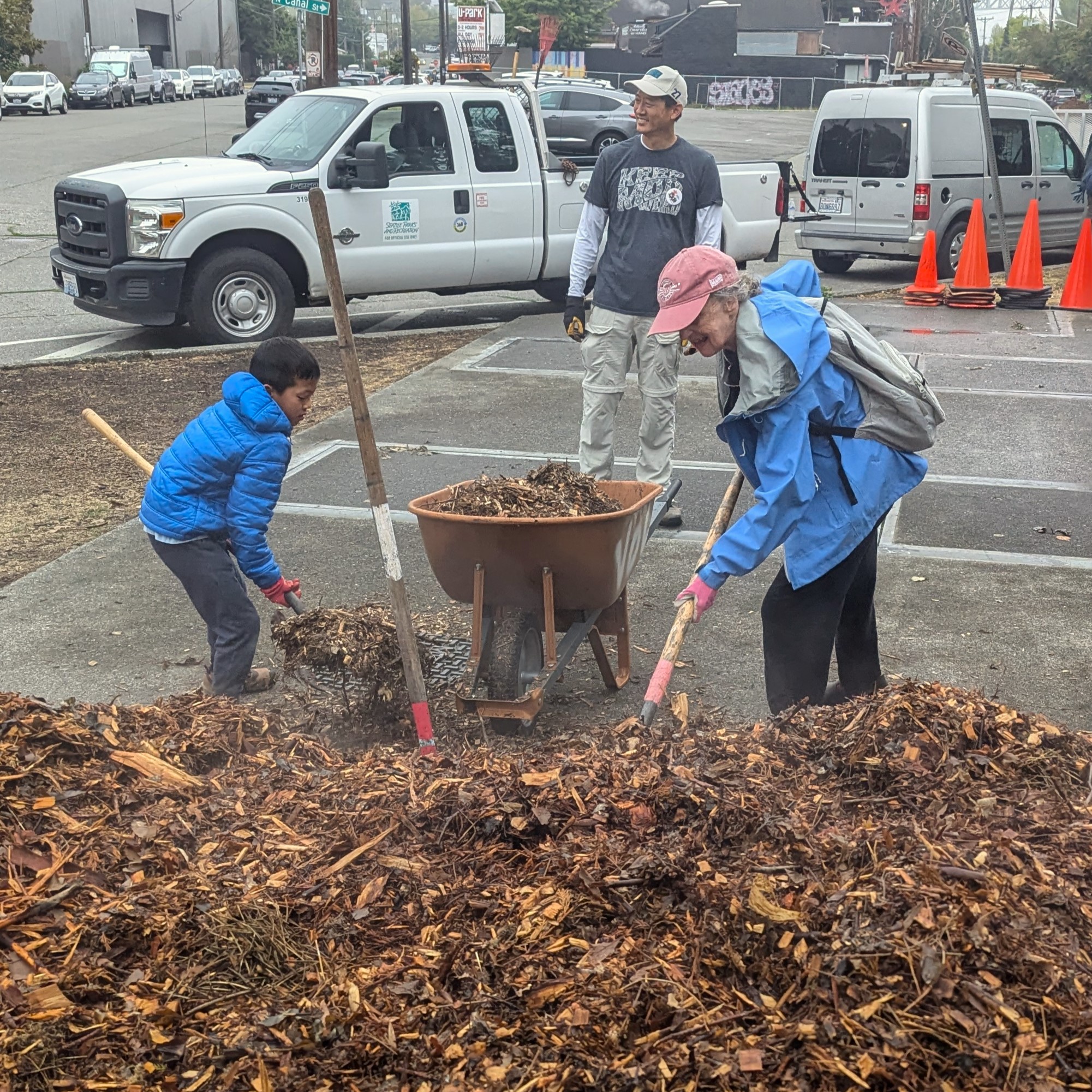 A woman and child work to shovel mulch from a pile into a wheelbarrow.