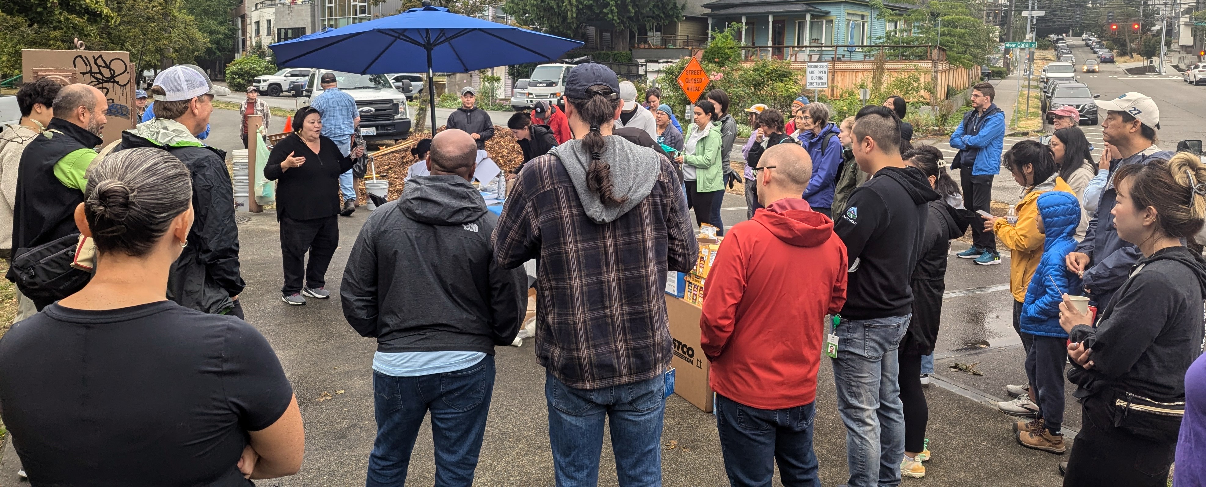 A large crowd of people in a circle listen to a woman standing under a blue umbrella give instructions