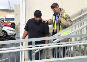 A man in a black tshirt and black cap helps a man in with a cane down a flight of stairs outside a building.