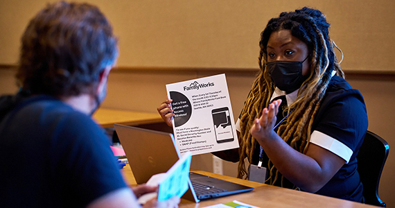 A white man and a Black woman sit across a table from each other, wearing masks. The woman has a laptop and holds up and explains a flyer about free cellphones.