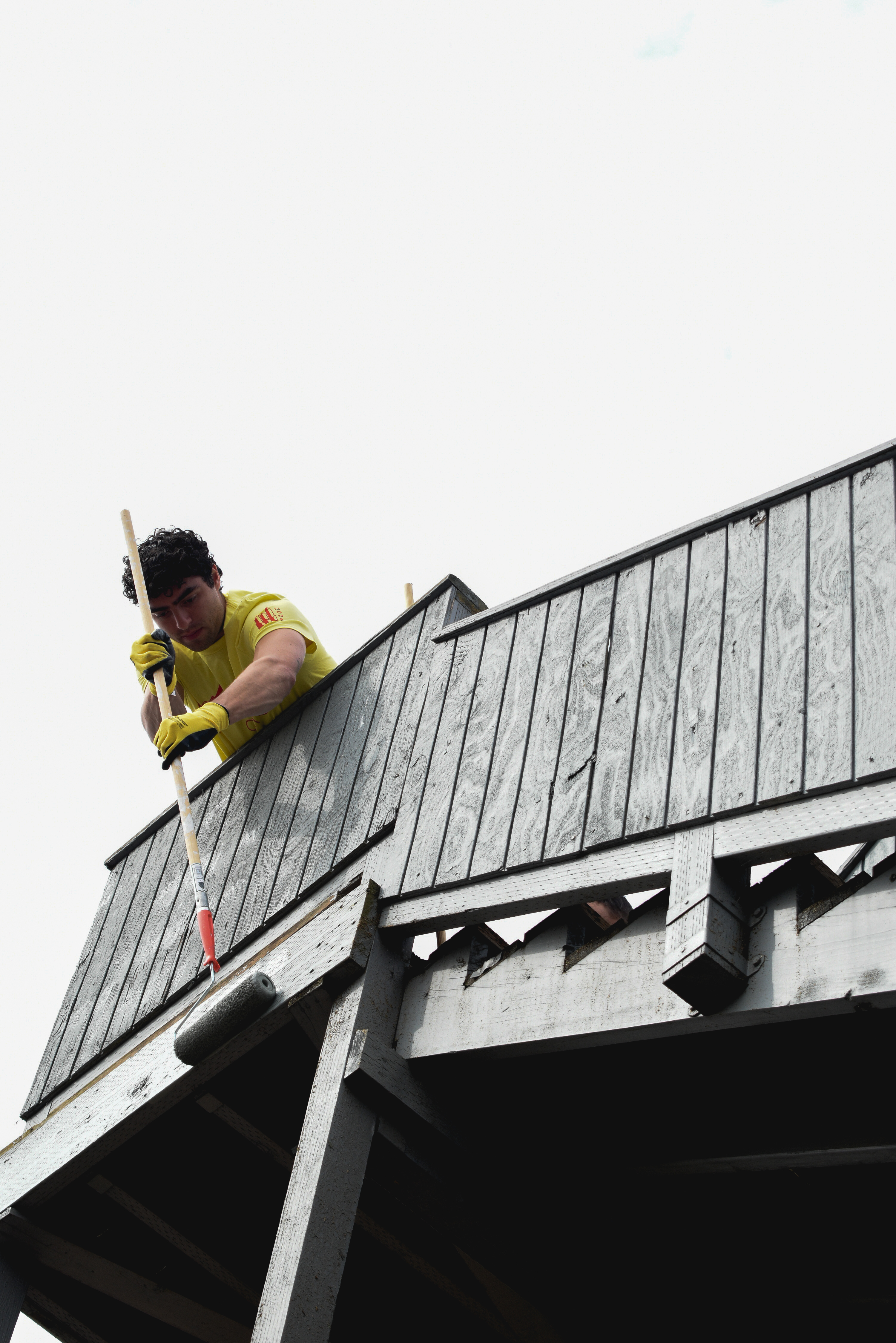 A volunteer in a yellow shirt leans over the railing of a wooden fire escape to apply a fresh coat of paint with a roller on a long pole.