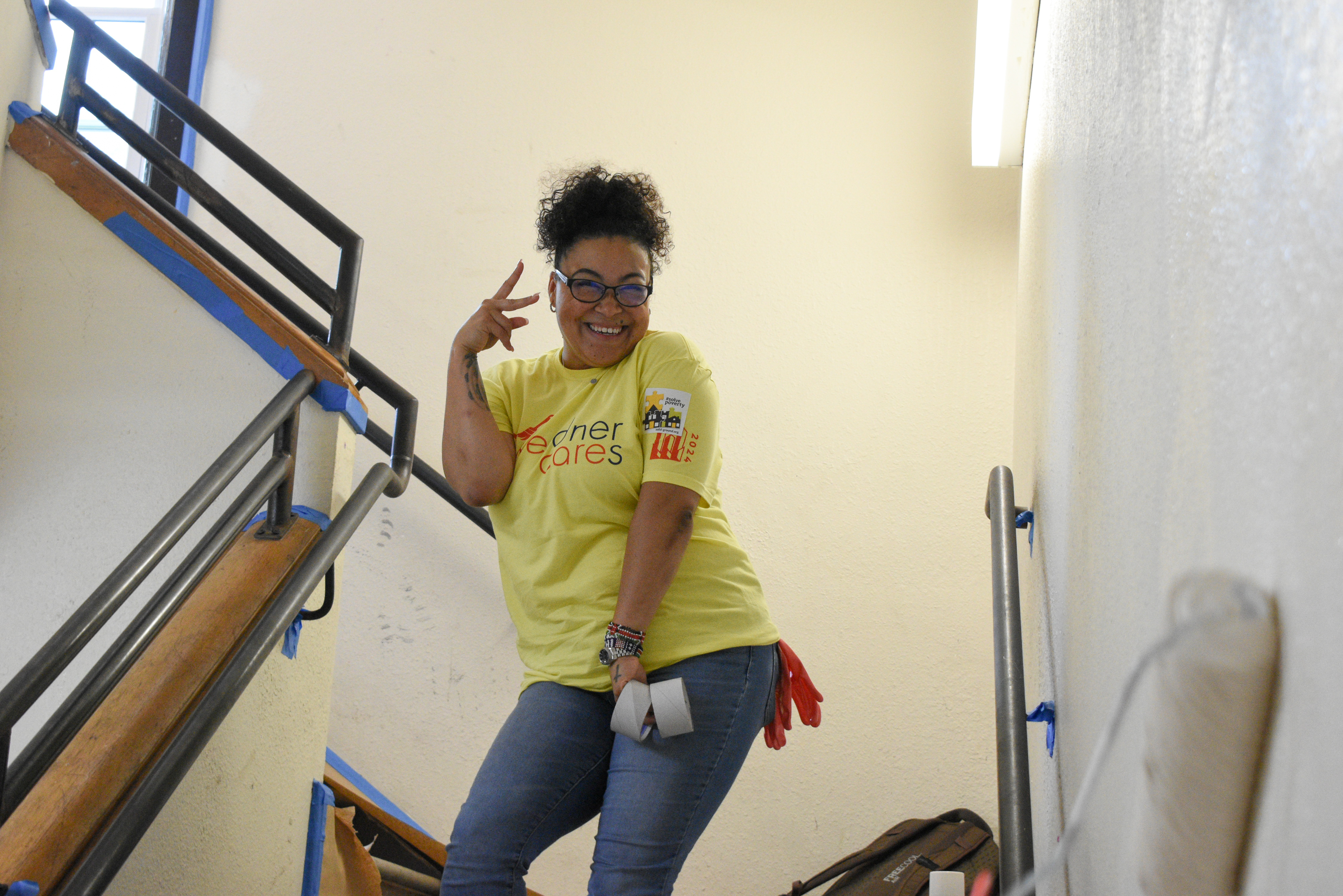A woman in a yellow t-shirt smiles and makes a peace sign with her fingers while standing in a freshly painted stairwell