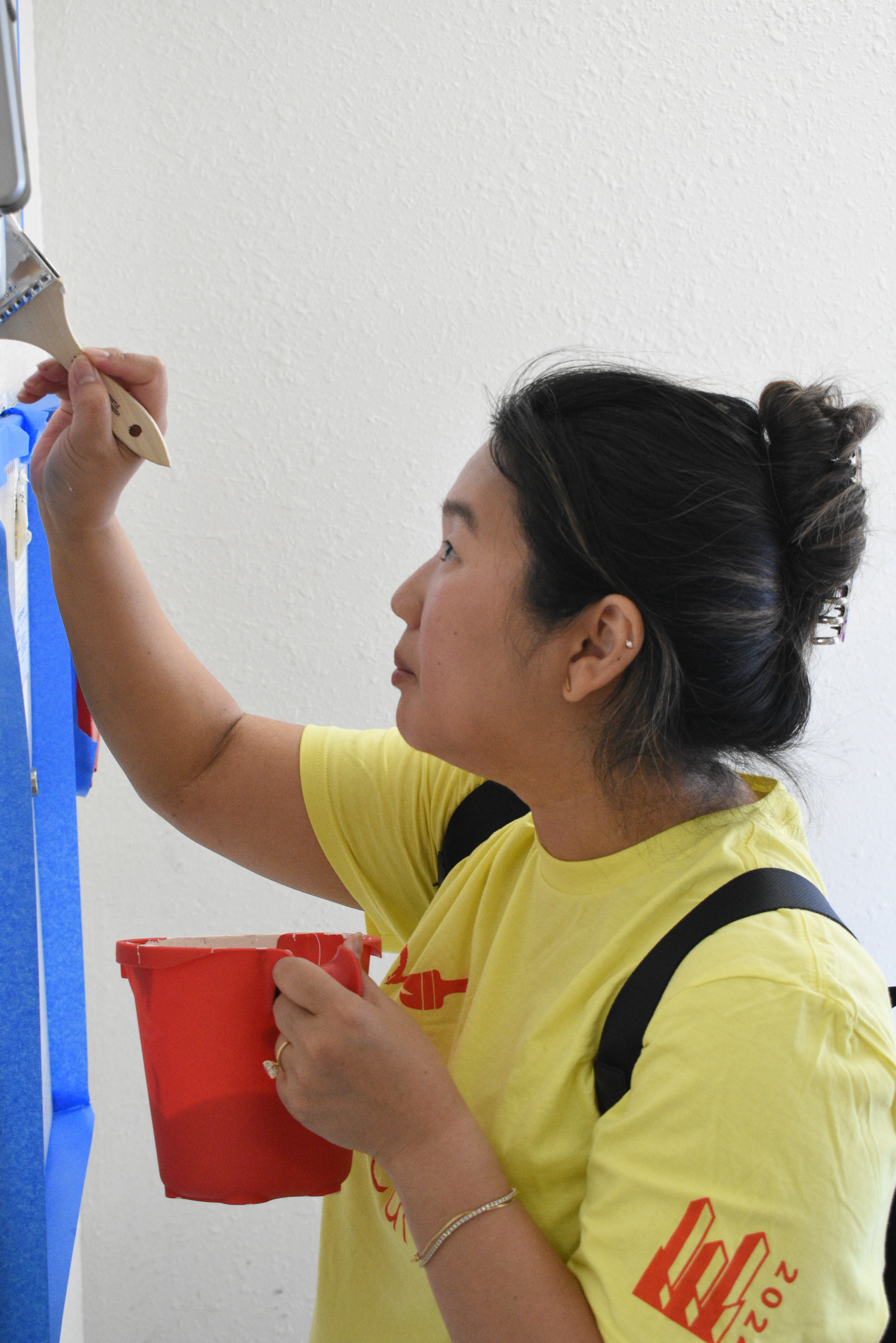 A woman in a yellow shirt holds a small red bucket of paint in one hand while painting a wall with the other.