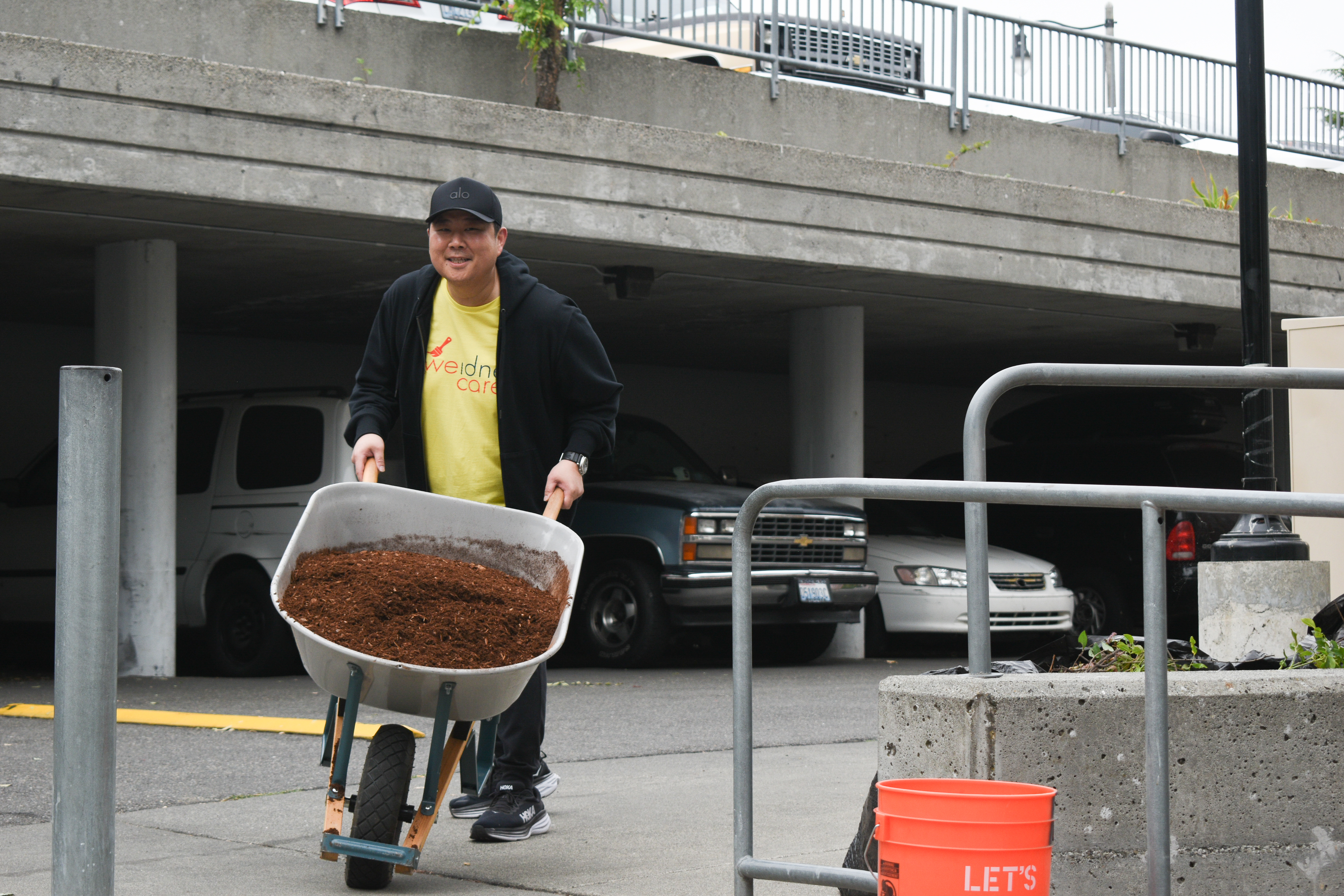 A man in a black jacket over a yellow t-shirt pushes a wheelbarrow full of mulch past a row of cars.