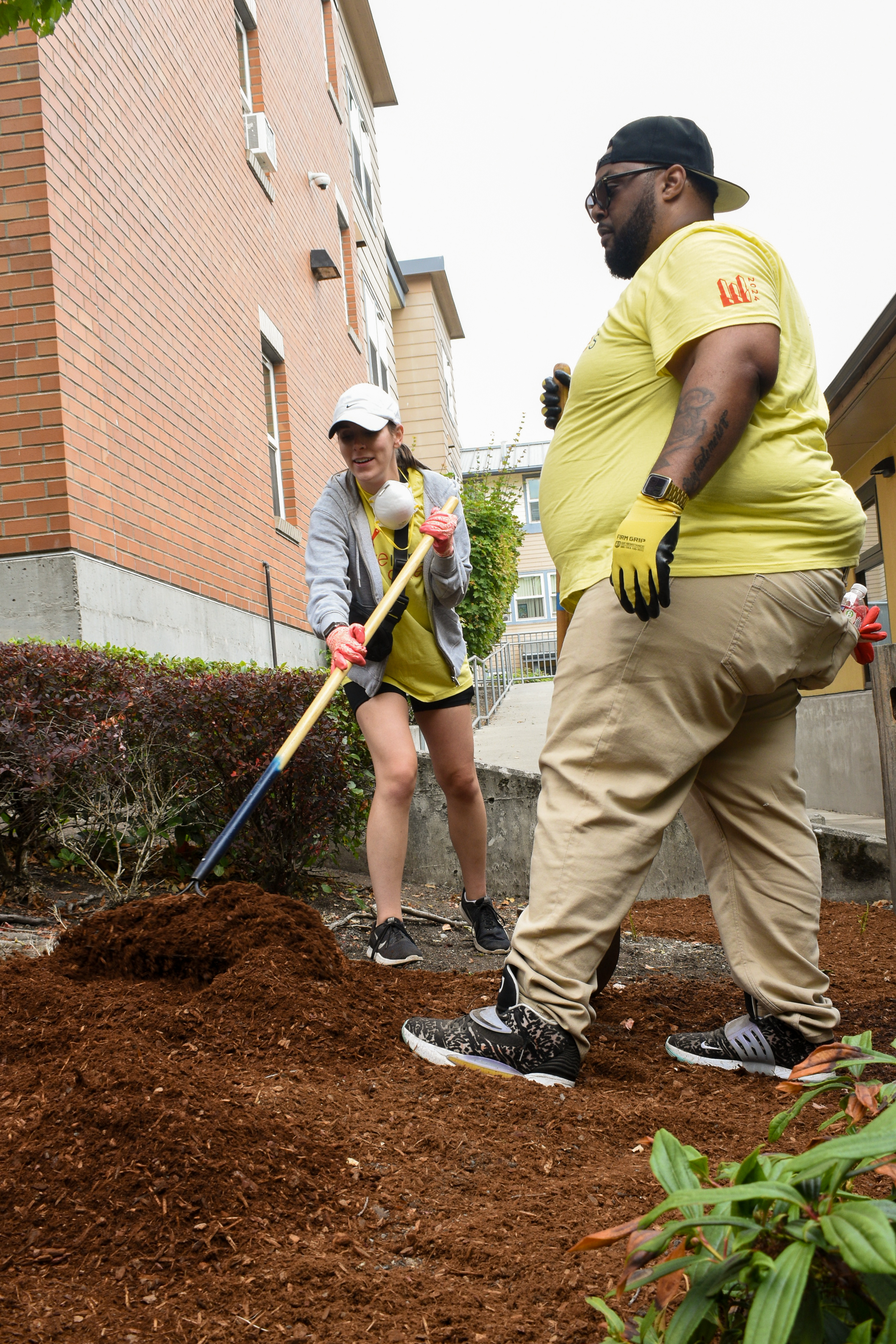 Two people in yellow shirts work to spread mulch near the corner of a brick building.