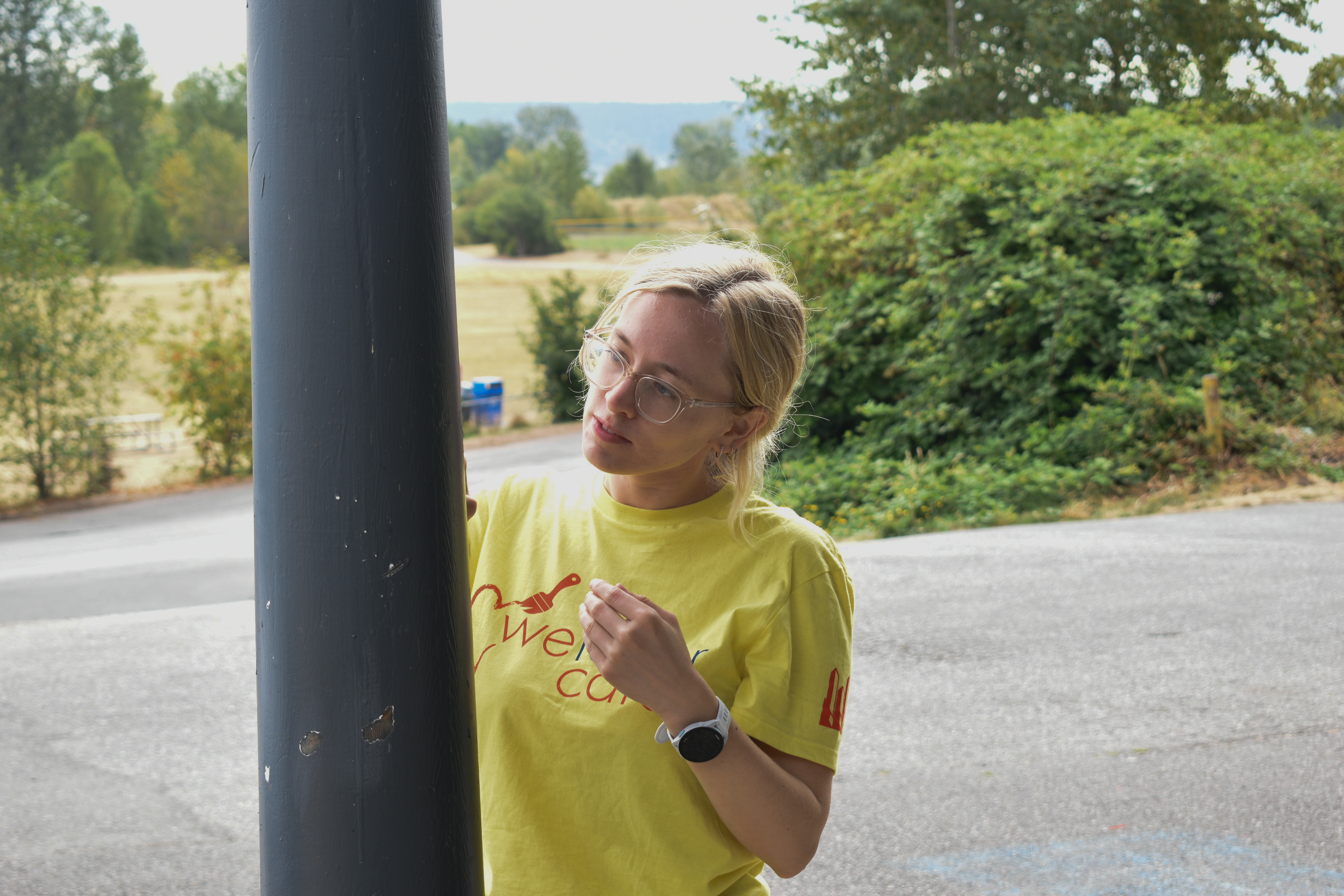 A volunteer in a yellow t-shirt carefully paints a gray column on the outside of a building.