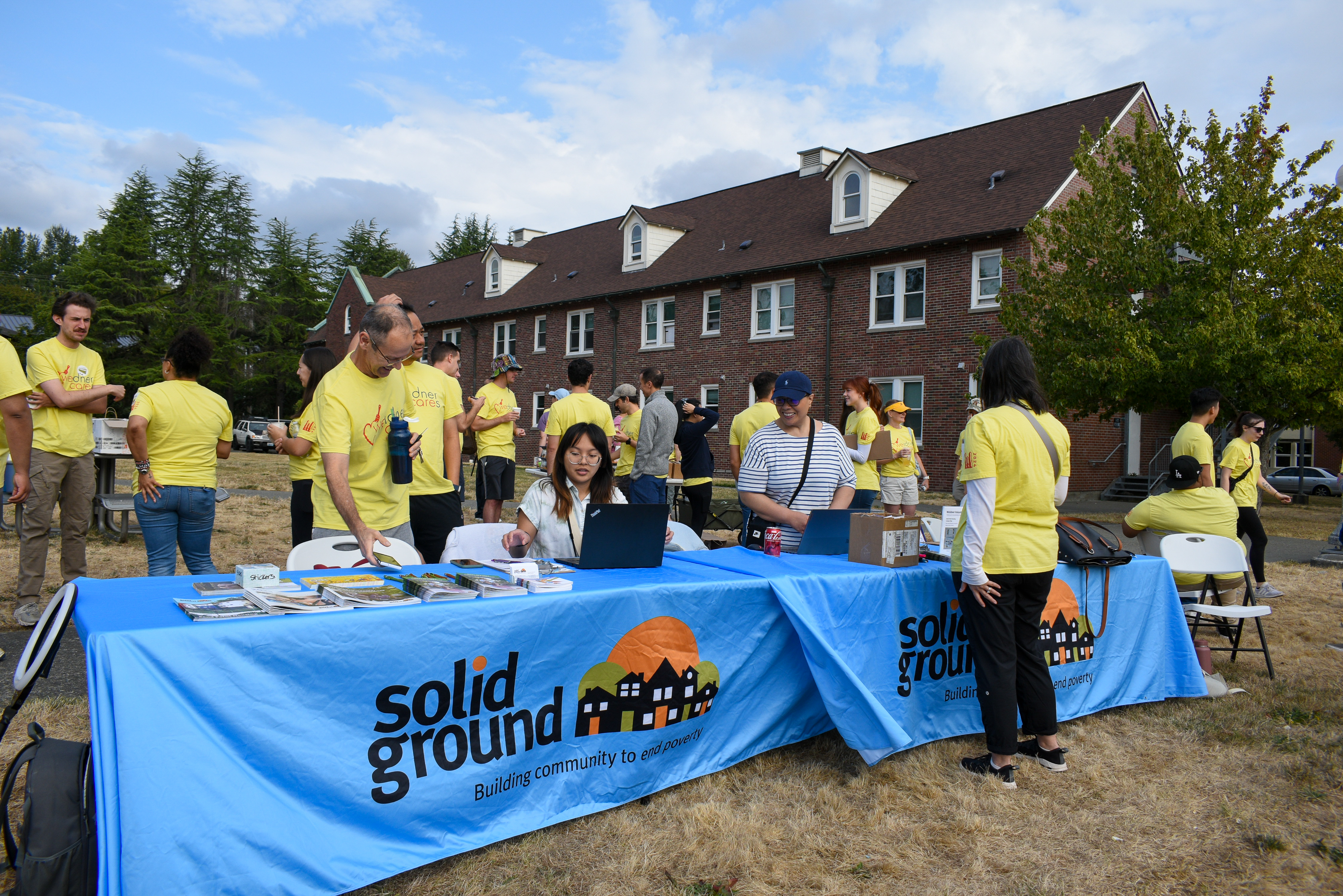 A group of people in yellow shirts congregate around a set of tables covered with blue table clothes with the Solid Ground logo.