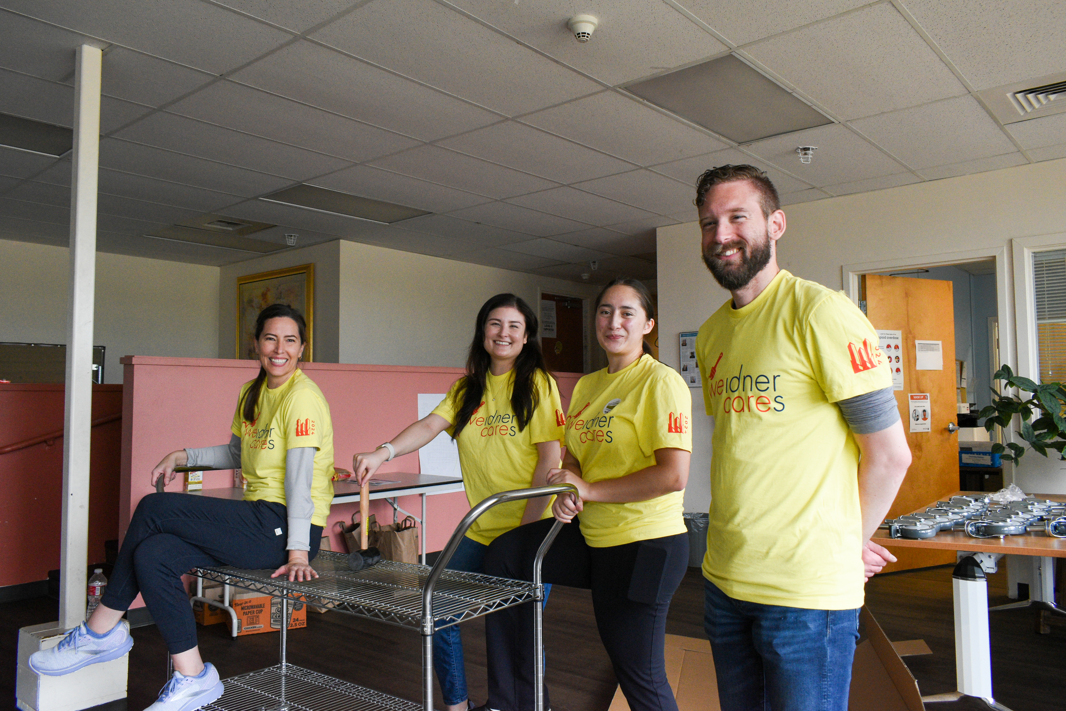 Three people in yellow t-shirts stand besides a metal car while a fourth person, also in a yellow shirt, sits on it with legs crossed