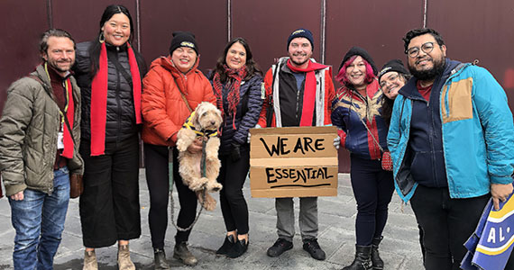 Eight people (and one dog!) wearing warm winter clothing pose for the camera. One man holds a sign on a piece of cardboard that reads WE ARE ESSTENTIAL.