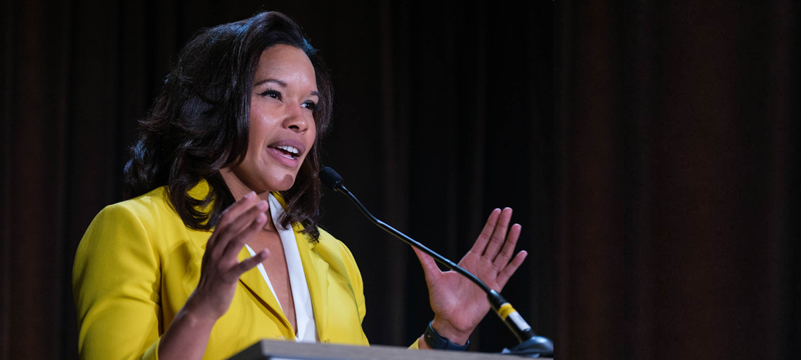 Woman with dark shoulder-length hair wearing a yellow blazer and white blouse gestures with her hands while speaking into the microphone.