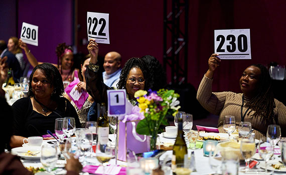 A group of smiling people in fancy dress raise bidder numbers high at a fancy dinner table.