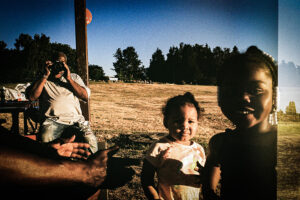 Photo of two little children smiling with a dried grass field behind them. One adult has his arms outstretched toward them; another is taking a photo of them from behind.
