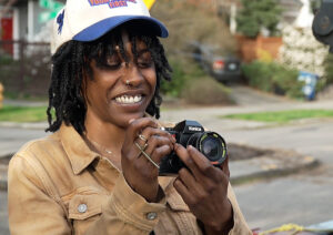 A young woman in a tan jacket and baseball cap smiles as she peers into her camera's window.