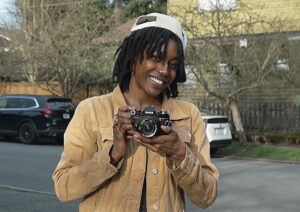 Portrait of a Black woman with short locs wearing a beige collared shirt, and holding a camera in her hands.