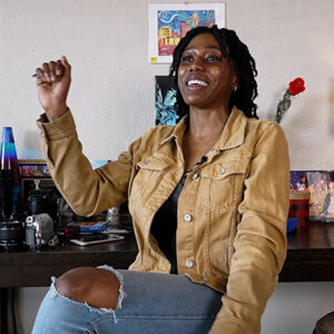 A young woman in a tan jacket sits inside her apartment, gesturing with her right fist raised.