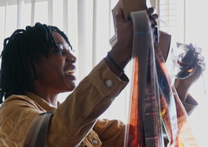 A young woman in a tan jacket smiles as she holds up strips of camera film.