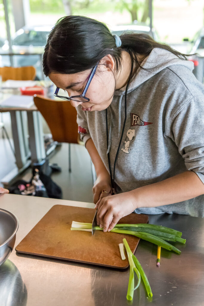A cooking class student carefully slices scallions on a cutting board.