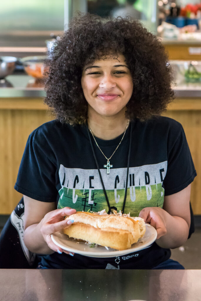 A young cooking class student smiles as she holds out a bahn mi sandwich on a plate.