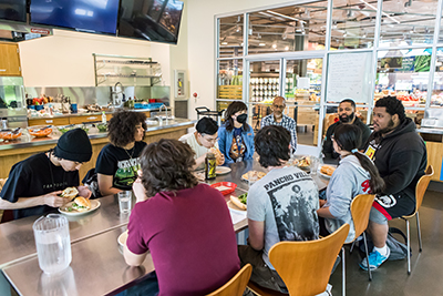 A group of cooking class students and instructors crowd around a table eating bahn mi sandwiches. A brightly lit grocery store is seen through floor-to-ceiling windows behind them.