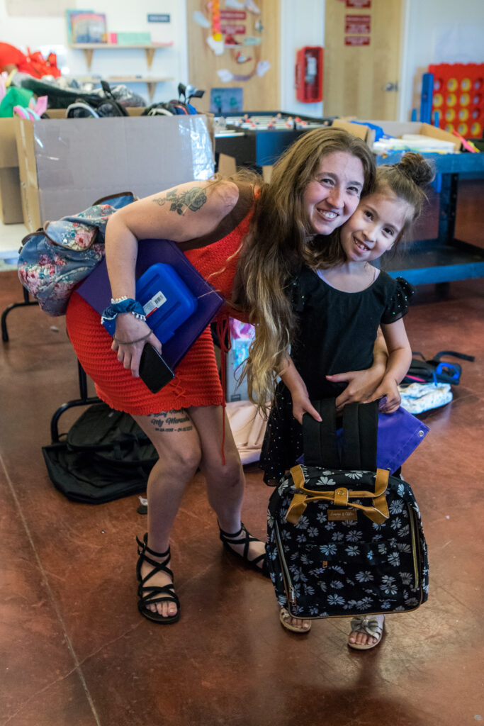 A smiling mother with long hair crouches down and empraces her young daughter, who is holding a new backpack.