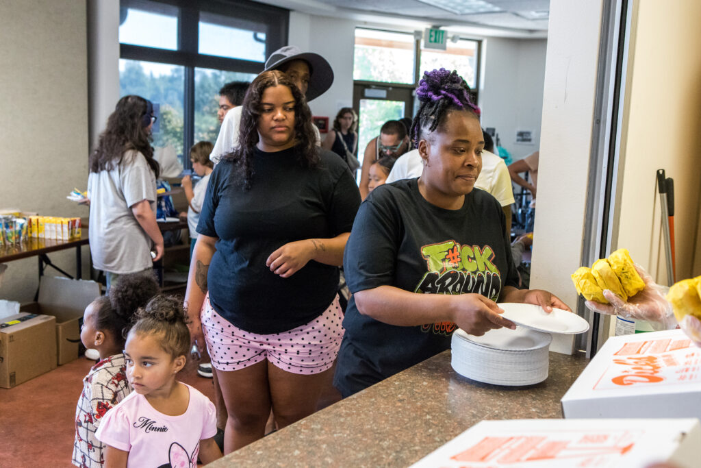 Two moms with kids are handed several hamburgers from a box labeled "Dick's."