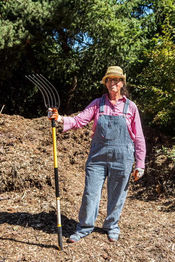A woman in overalls holds a pitchfork in front of a pile of mulch.