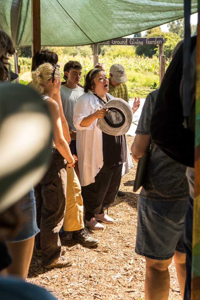 A woman takes off her hat to address a group gathered under a tarp at a farm.