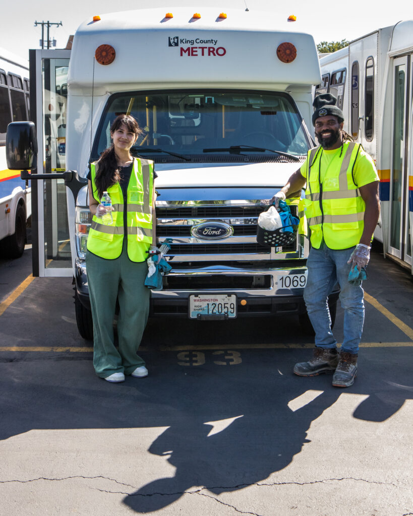 Two people holding cleaning supplies and wearing yellow safety vests pose in front of a paratransit bus.