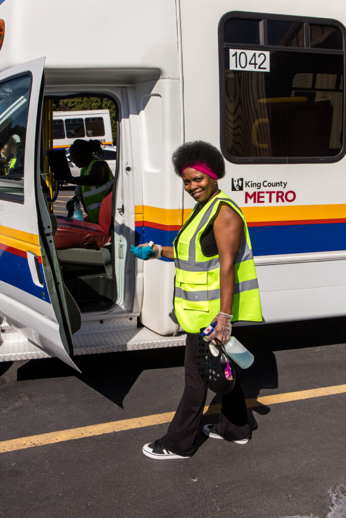 A woman wearing a yellow safety vest and holding a spray bottle stands next to the open cab of a paratransit bus.