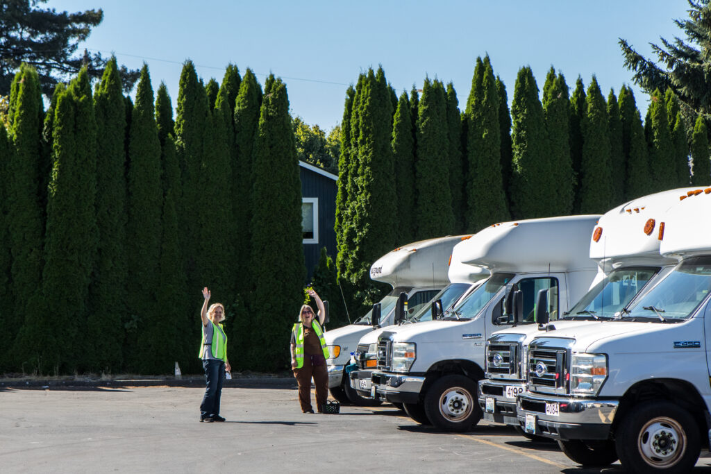 Two people in yellow safety vests wave at the camera from in front of a row of paratransit buses.