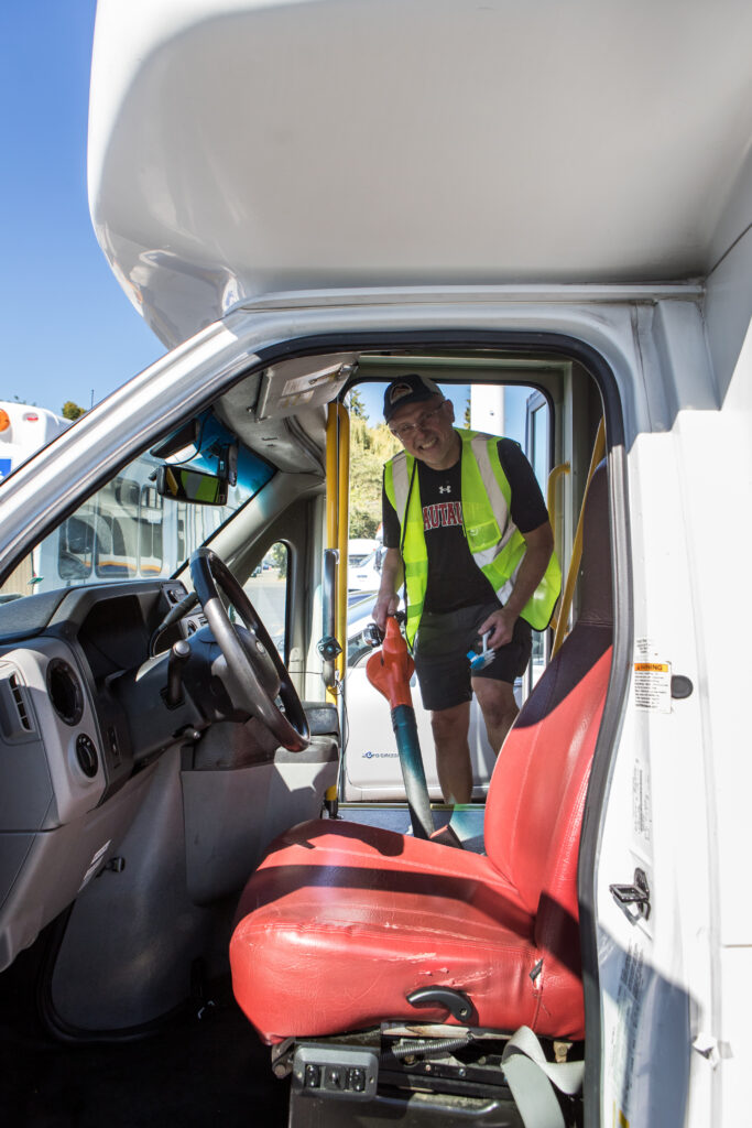 A man in a yellow safety vest and holding a blower cleans the cab of a paratransit bus.