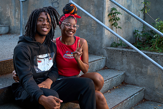 A young couple wearing black and red pose on a concrete staircase. They both smile, and she makes a peace sign.