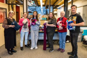 Six people pose for a photo while holding knitted items.