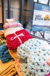 Several knitted items stacked in front of a window with the Solid Ground sign visible outside.